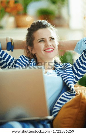 happy 40 years old housewife in blue blouse and striped jacket sitting on divan with laptop in the modern house in sunny day.