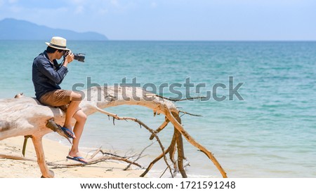 Asian man photographer and tourist wearing hat taking photo on dead pine tree on tropical island beach. Relaxing holiday or vacation travel in summer season. Summertime landscape photography concept