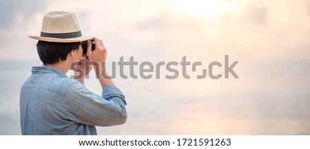 Asian man tourist and photographer wearing hat taking photo on tropical island beach during sunset. Relaxing holiday or vacation travel in summer season. Summertime landscape photography concept
