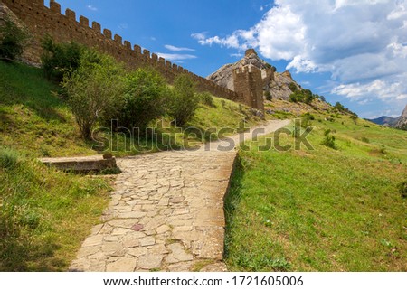 An ancient fortress on top of a mountain and path along the fortification wall in sunny day. Genoese fortress, Sudak, Crimea. Historic old building. Monument of architecture.  Royalty-Free Stock Photo #1721605006