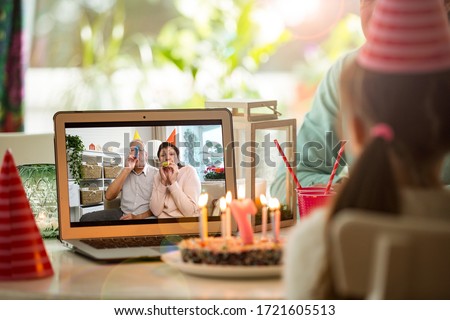 Happy little girl celebrating birthday at home with parents and grand parents on video call. Laptop with senior couple online, cake with candles on table.