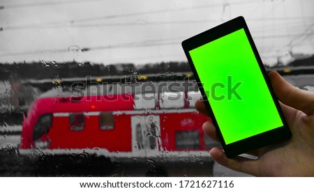 Smartphone with a green screen for effects or text with advertising. Against the background of a red passenger train and a wet window with water drops after the rain. Grey sky with clouds. Template. 