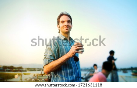 A man holding a can of beer or soda in his hand smiling positively, there is a ray of sunset light behind him. There are blurred people and a lake or river behind him Royalty-Free Stock Photo #1721631118