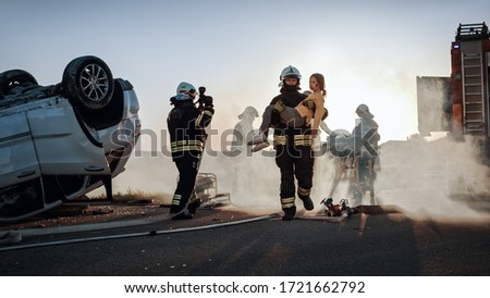 Brave Firefighter Carries Injured Young Girl to Safety where She Reunited with Her Loving Mother. In the Background Car Crash Traffic Accident Courageous Paramedics and Firemen Save Lives