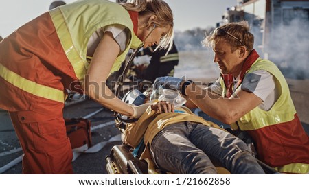 On the Car Crash Traffic Accident Scene: Paramedics Saving Life of a Traffic Accident Victim who is Lying on Stretchers. They Listen To a Heartbeat, Apply Oxygen Mask and Give First Aid Help Royalty-Free Stock Photo #1721662858