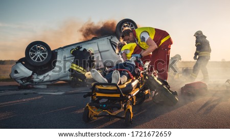 On the Car Crash Traffic Accident Scene: Paramedics Save Life of a Female Victim Lying on Stretchers. They Listen To a Heartbeat, Apply Oxygen Mask and Give First Aid. Firefighters Extinguish Fire Royalty-Free Stock Photo #1721672659