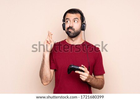 Man playing with a video game controller over isolated wall with fingers crossing and wishing the best