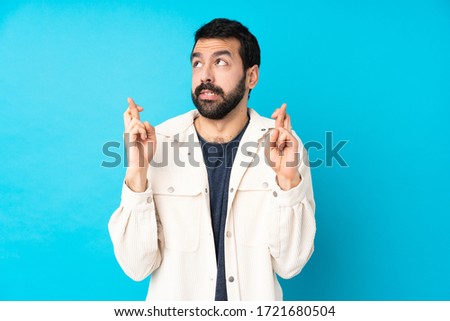 Young handsome man with white corduroy jacket over isolated blue background with fingers crossing and wishing the best