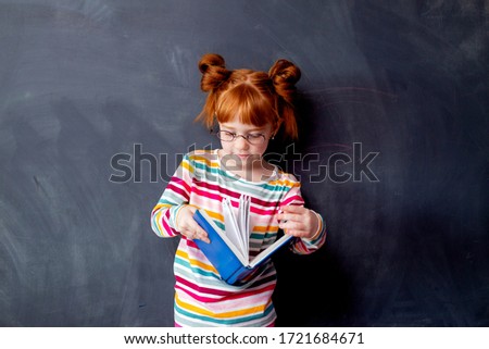 little girl with red hair is smiling standing in the background of the blackboard, holding a book. the education of the child, space for text