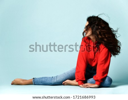 Flirting curly brunette adult woman in smoked blue glasses, red sweater and blue jeans jeggings is sitting posing on the floor looking aside at copy space over blue background