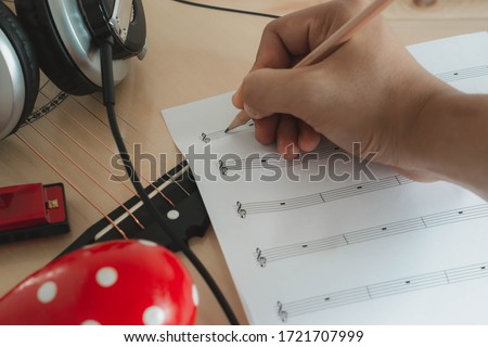 Music instruments concept background. Close up musician writing note on the paper for create song with the group of musical tools the guitar, headphone, maracas & harmonica in the morning time.
