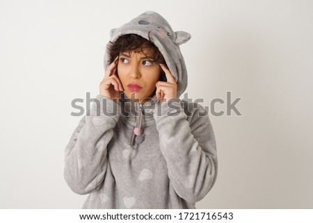 Dreamy Caucasian wearing pajama female with thoughtful expression, looks away, keeps hand near face, thinks about something pleasant, poses against gray wall.