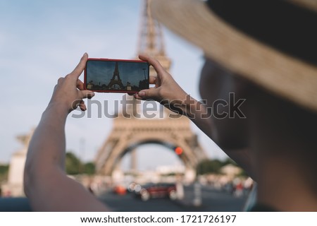 Anonymous lady in hat using smartphone to shoot exterior of Eiffel Tower while spending time on street of Paris on sunny day in France