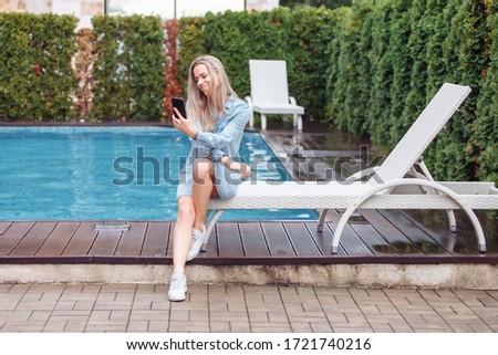 Well-groomed beautiful young modern woman makes video call to her work colleagues while she is on vacation. Cute girl is sitting in her own garden with pool on patio. Long distance communication