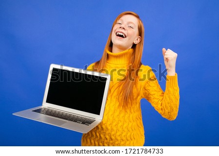 happy red-haired teenager shows a laptop display with a layout for advertising on a blue studio background
