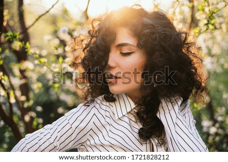 Beautiful brown girl enjoying nature in blooming garden. Outdoor portrait of a Young Woman with spring flowers. Romantic fashion model in blossom flowers portrait. Girl wearing stylish clothes. 
