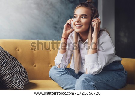 Young beautiful woman listens to music in headphones. Girl in white shirt sits on couch enjoys streaming audio, podcasts
