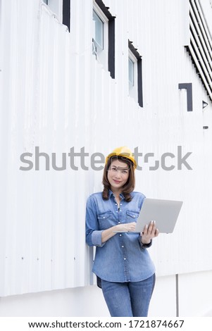 Beautiful Asian female engineer in jeans dress wearing yellow safety hard hat using laptop computer notebook doing job at construction site outside office. Idea for modern working woman.