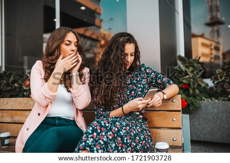 Image of curious woman spying and peeping at smartphone of her friend, while talking on mobile phone and sitting on the bench in the city street
