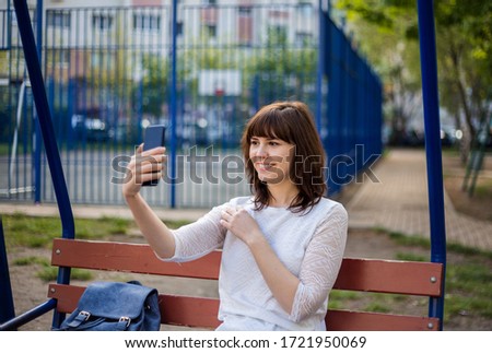 A young girl takes photos of herself on the phone on the street. A cute girl is sitting on a bench and smiling, holding a phone in her hands. A girl in a white jacket and jeans.