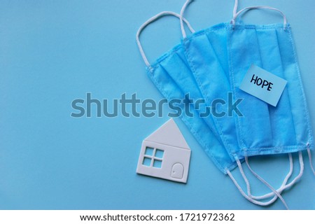 A three-layer medical mask with rubber ear straps, white paper home and blue stiker with the word hope. The concept of medicine. Hope. Stay home, stay safe.