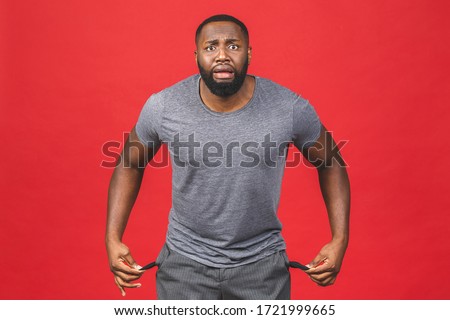 Portrait of frustrated worried african american black man with beard in casual turning out empty pockets showing I have no money gesture, bankrupt. Isolated on red background. Royalty-Free Stock Photo #1721999665