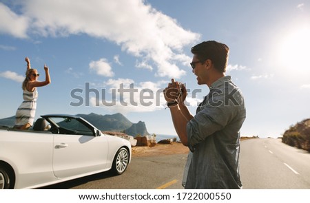 Man taking photos of a woman in a convertible car with a camera on highway. Couple doing a photo shoot on road trip.