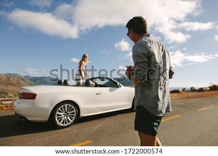 Man with camera on the highway with woman standing inside a open car. Couple having a photo shoot on their road trip.