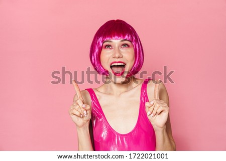Image of excited girl wearing wig expressing surprise and pointing fingers upward isolated over pink background