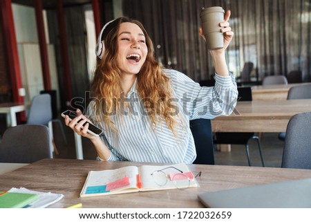 Image of excited young woman singing while using cellphone and wireless headphones at classroom