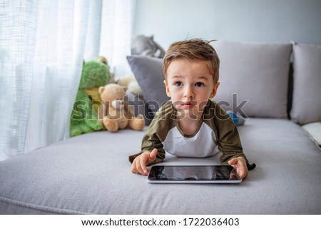 Adorable toddler boy laying on the sofa and playing with tablet pc at home, indoors. Child with tablet computer. Little boy using smart tablet at home. Little boy with touch pad, early learning