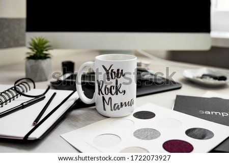 Motivational sign on a white cup of a mother's working place. Monochromatic office stuff and black stationery with a computer on the background. 