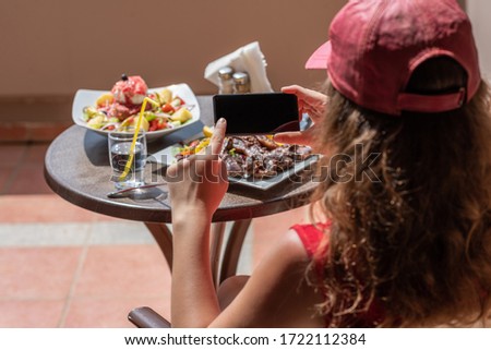 Woman blogger taking a picture on mobile phone  plate with lamb potato and salad on the table outside on the balcony. Top view.