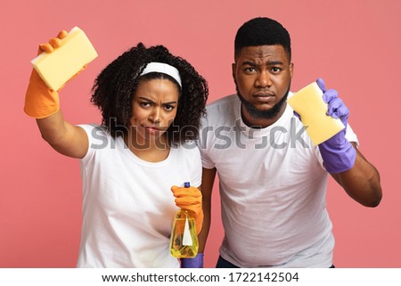 Accurate Black Couple Holding Cleaning Tools And Looking Suspiciosly At Camera, Standing Together Over Pink Studio Background, Closeup