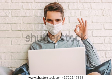 Young hipster man in protective face mask showing okay sign, while having online video call at home. Happy man in medical face mask sit on couch, talk on virtual web chat. Quarantine communication