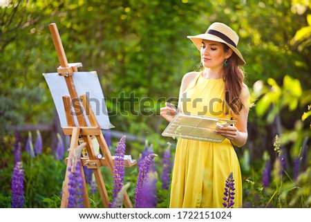 A girl artist in a yellow dress and hat draws a lupine field in the open air. Easel box with canvas, oil paint, palette and artist 's tools.