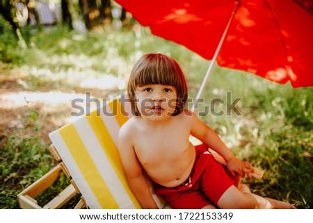 Funny little boy in red shorts lying on the striped yellow and white deck chair on the background of red sun umbrella on the nature in summer park