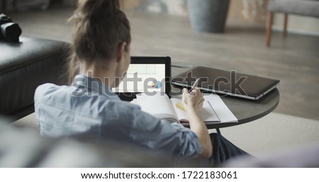 Girl sitting on sofa and using digital tablet computer in home indoors, making notes. Young woman shopping online. Freelance female model working out of office. Back right view  Royalty-Free Stock Photo #1722183061