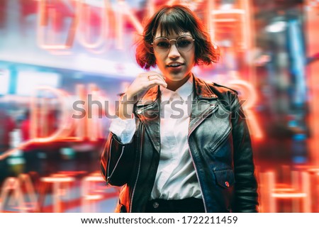 Half length portrait of attractive Caucasian party girl posing near neon wall during night walk in New York, beautiful female tourist in trendy streetwear and stylish spectacles for vision correction Royalty-Free Stock Photo #1722211459