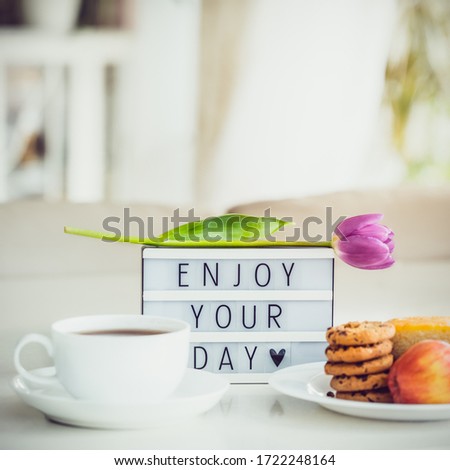 Good morning concept. Romantic breakfast - cup of hot drink, cookies, lightbox with message Enjoy your day and tulip flower on marble table with light interior view. Close up. Square card. Copy space