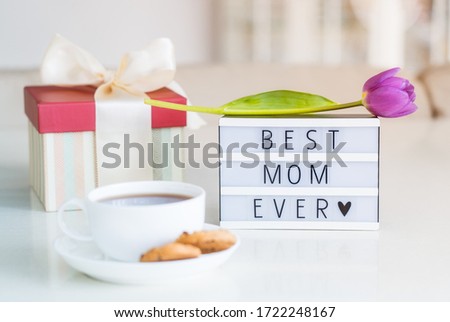 Mother's day background. Morning suprise - cup of tea with cookies, lightbox with words Best mom ever and tulip flower on it standing on the marble table with light interior view. Close up. Copy space