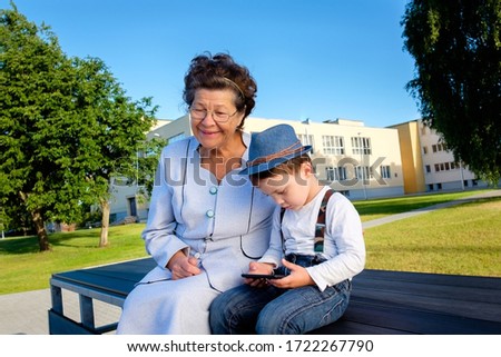Happy grandmother with glasses sitting on bench at park while grandson playing games, reading story, watching cartoon with smartphone in hands at city park. Concept of happy family day, technology