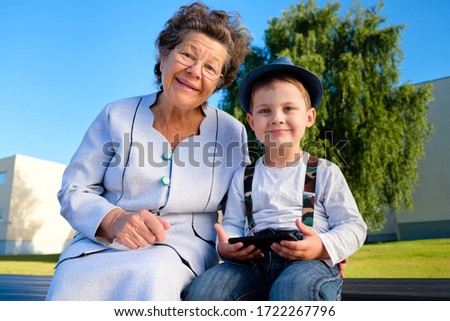 Happy smiling grandmother with glasses and grandson wearing jeans with suspenders fedora hat sitting on bench with smartphone at city park, looking at camera. Concept of happy family, emotions