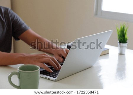 A man uses a laptop at home while sitting at a wooden table. Man hands typing on a notebook keyboard. The concept of young people working at mobile devices. The background is wide blurry window.