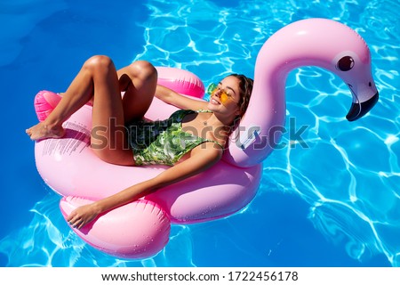 Young smiling fitted girl in bikini relax chilling on pink inflatable flamingo in swimming pool. Attractive woman in swimsuit lies in a sun on tropical vacation. Pretty female sunbathing at resort.