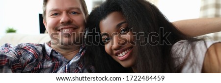 Portrait of cheerful mixed race couple taking happy selfie on sofa indoors. Man and latino woman widely smiling on camera. Multinational and biracial family concept