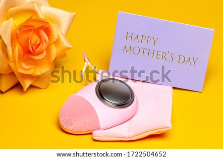 Mothers day greeting card. Close-up of a modern electric face cleansing brush a beautiful yellow blossom of flower and a tag with inscription Happy mothers day on yellow background. Beauty for the mom