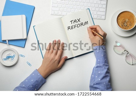 Woman writing phrase TAX REFORM in notebook at white table, top view