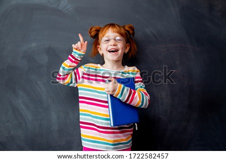 little girl with red hair is smiling standing in the background of the blackboard, holding a book. the education of the child, space for text