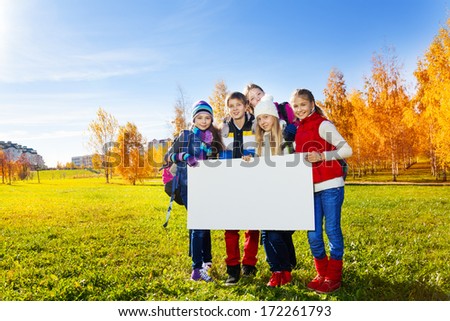 Group of happy teen kids standing in autumn park with blank white board poster for adding text